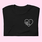 Flat lay of a black unisex t-shirt folded neatly on a pink background. A small white left-chest print reads “I’ve heard enough from old white men,” surrounded by a simple heart outline.