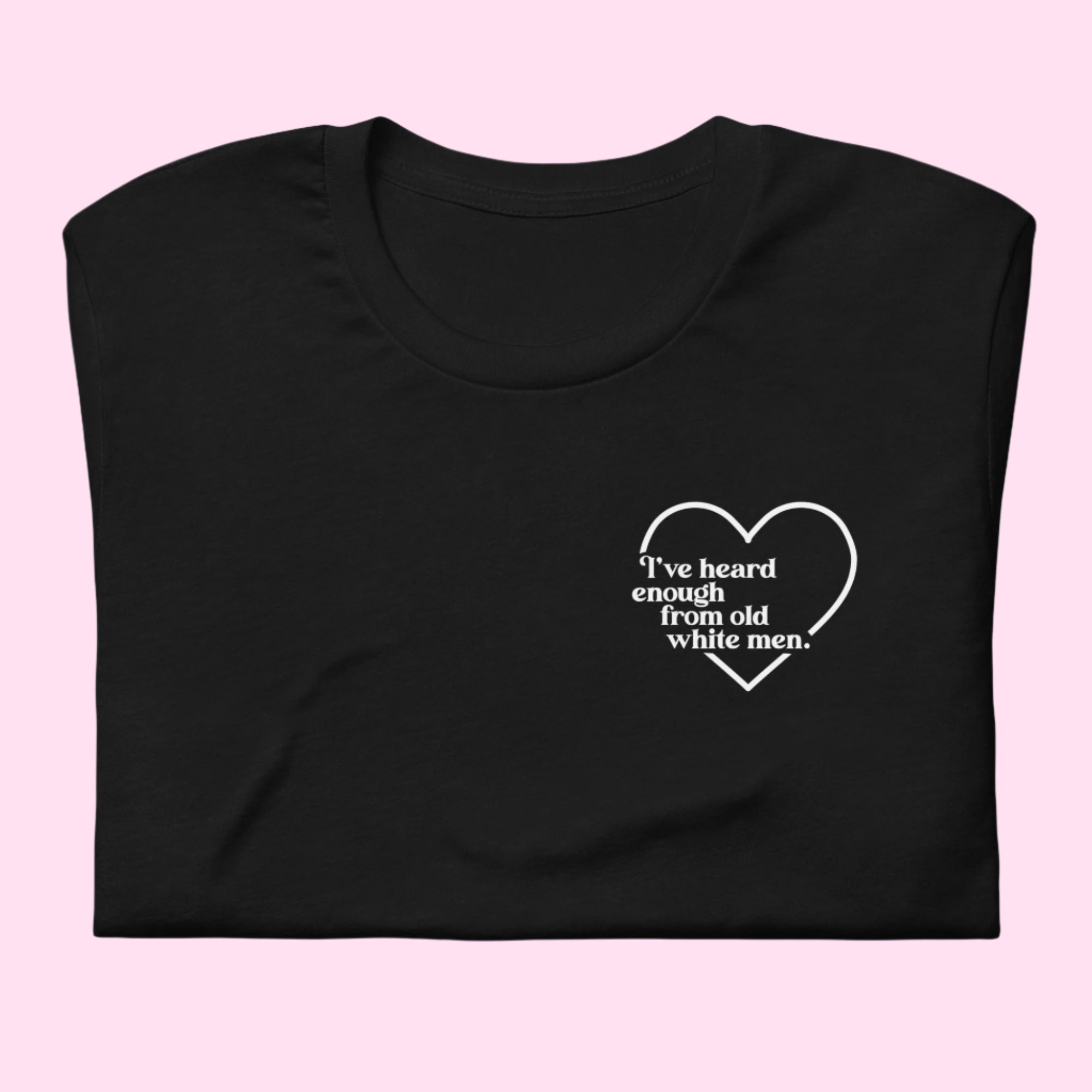 Flat lay of a black unisex t-shirt folded neatly on a pink background. A small white left-chest print reads “I’ve heard enough from old white men,” surrounded by a simple heart outline.