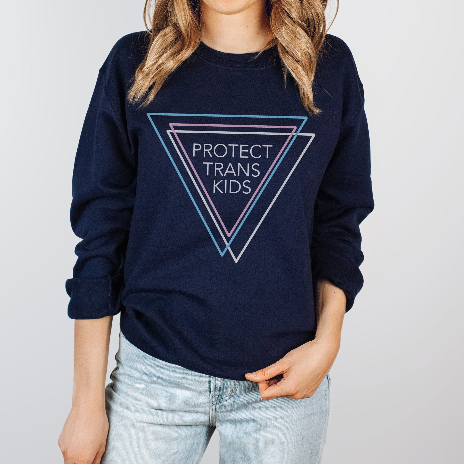 Woman wearing a navy blue sweatshirt that says 'Protect Trans Kids' with three triangles in blue, pink, and white representing the transgender flag.