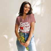 A woman smiling and holding a bouquet of flowers while wearing a mauve-colored relaxed v-neck t-shirt. The shirt reads “Care About Other People” in bold text, with the “O” in “Other” replaced by a small heart. The soft v-neck and relaxed fit create a casual, feel-good look.
