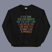 Flat lay of a black sweatshirt on a neutral background. The sweatshirt features a bold, multicolored quote across the chest that reads: "If you think you love freedom but you don't care if we are all free then what you actually love is privilege."