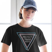 A woman wearing a black crop top that reads "Protect Trans Kids" in bold white letters, with three triangles in blue, pink, and white representing the transgender flag. She pairs the top with high-waisted jeans and a casual hat, standing with a relaxed, confident stance.