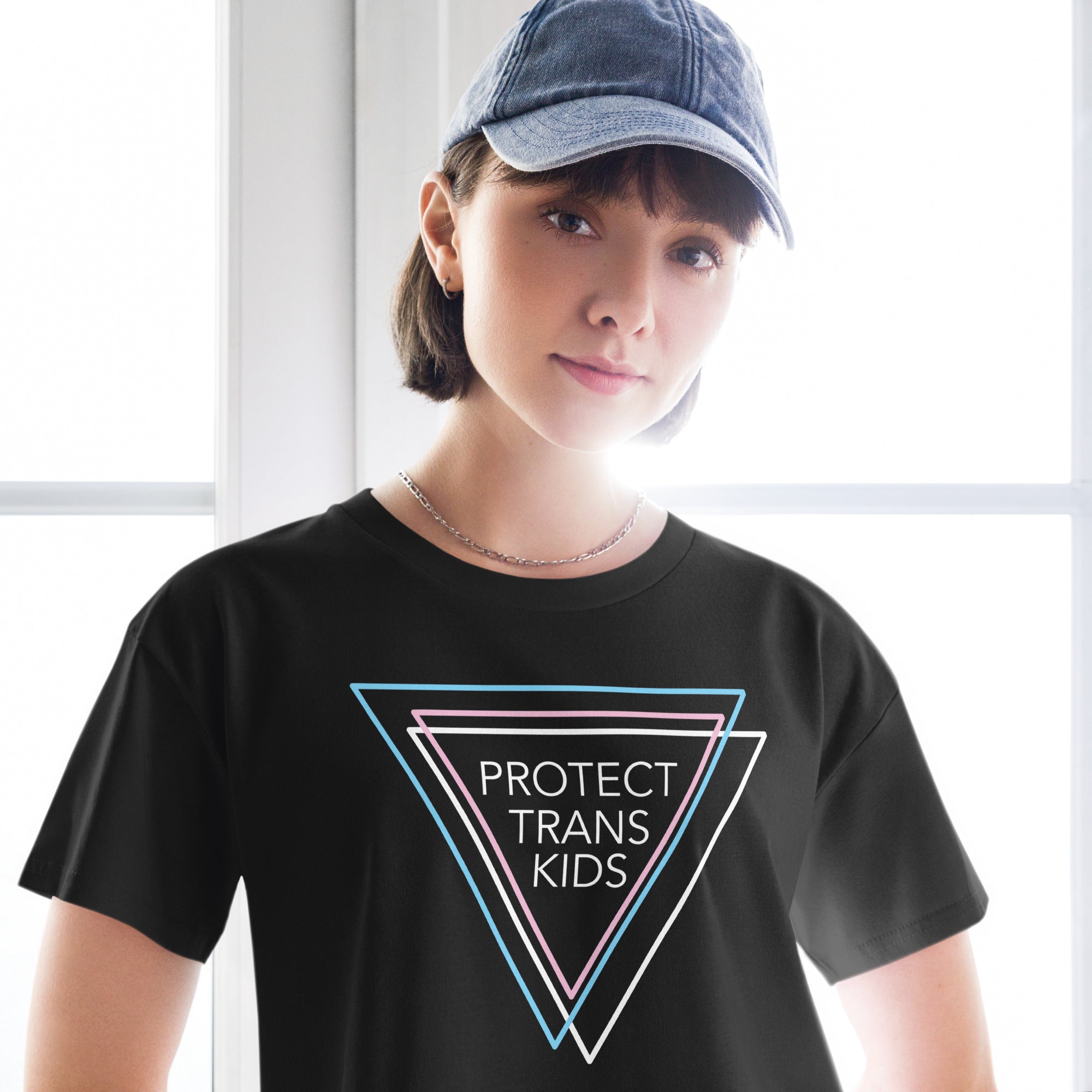 A woman wearing a black crop top that reads "Protect Trans Kids" in bold white letters, with three triangles in blue, pink, and white representing the transgender flag. She pairs the top with high-waisted jeans and a casual hat, standing with a relaxed, confident stance.