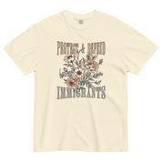 Ivory-colored unisex Comfort Colors t-shirt with the phrase “Protect & Defend Immigrants” in lightweight, uppercase font, surrounding a large, detailed flower graphic in the center. The design is bold yet soft, printed on a heavyweight cotton shirt with a relaxed fit.