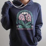 Woman wearing a heather navy hoodie with white drawstrings that reads “Immigrants deserve human rights. Stop funding cruelty.” in green and pink text with a floral design, up close shot showing soft fleece fabric and clear lettering.