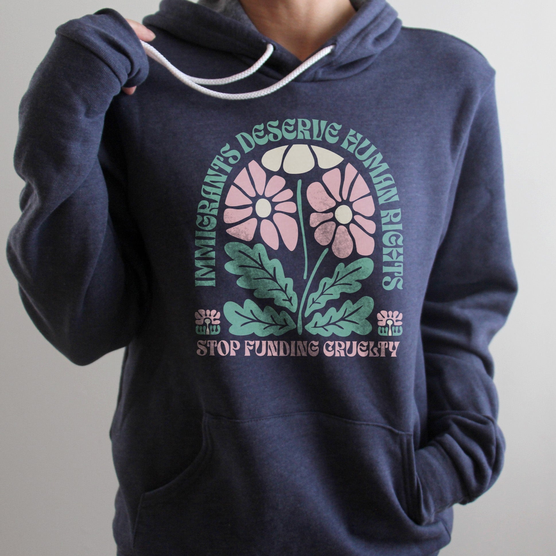 Woman wearing a heather navy hoodie with white drawstrings that reads “Immigrants deserve human rights. Stop funding cruelty.” in green and pink text with a floral design, up close shot showing soft fleece fabric and clear lettering.