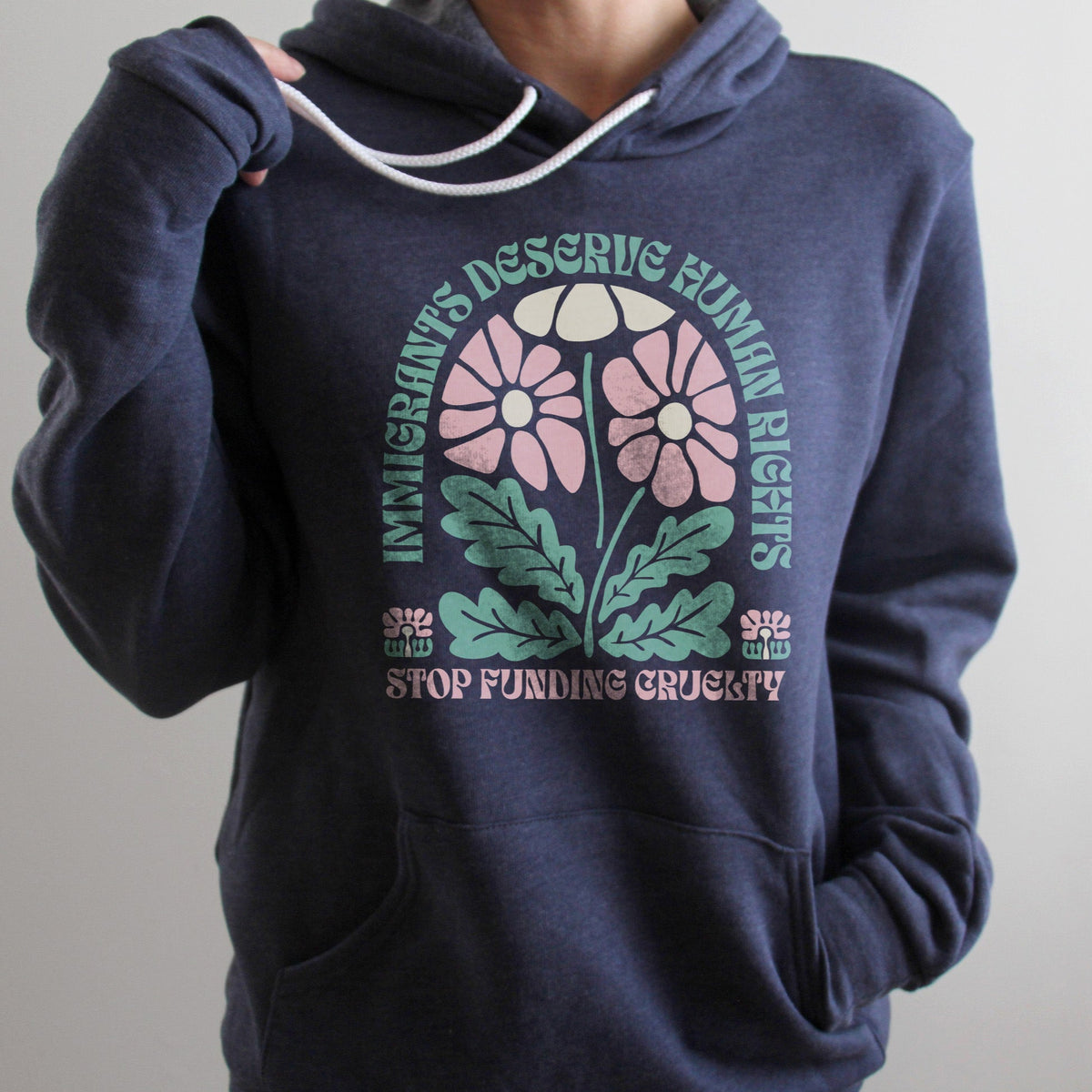 Woman wearing a heather navy hoodie with white drawstrings that reads “Immigrants deserve human rights. Stop funding cruelty.” in green and pink text with a floral design, up close shot showing soft fleece fabric and clear lettering.