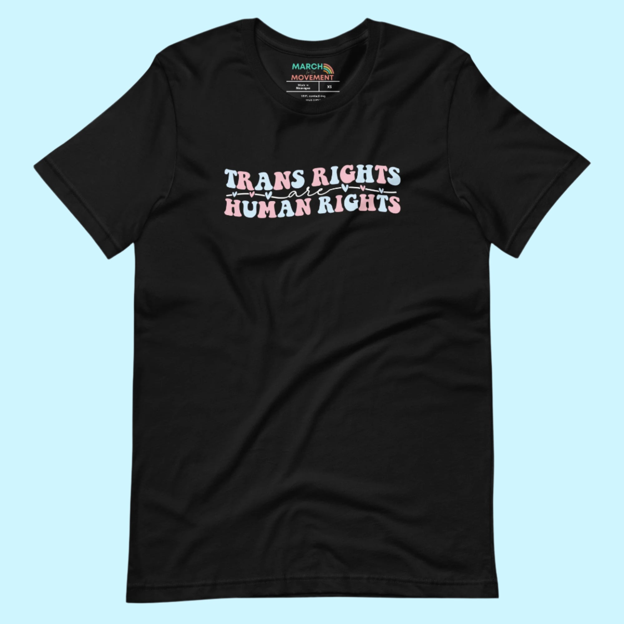 A black unisex t-shirt on a light blue background. The shirt features the phrase “Trans Rights Are Human Rights” written in the colors of the transgender flag—blue, white, and pink—with small heart accents throughout the design.