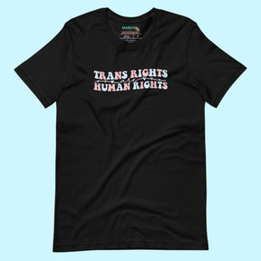 A black unisex t-shirt on a light blue background. The shirt features the phrase “Trans Rights Are Human Rights” written in the colors of the transgender flag—blue, white, and pink—with small heart accents throughout the design.