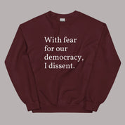 Flat lay of a maroon sweatshirt on a neutral background. The sweatshirt features white serif text across the chest that reads: “With fear for our democracy, I dissent.”