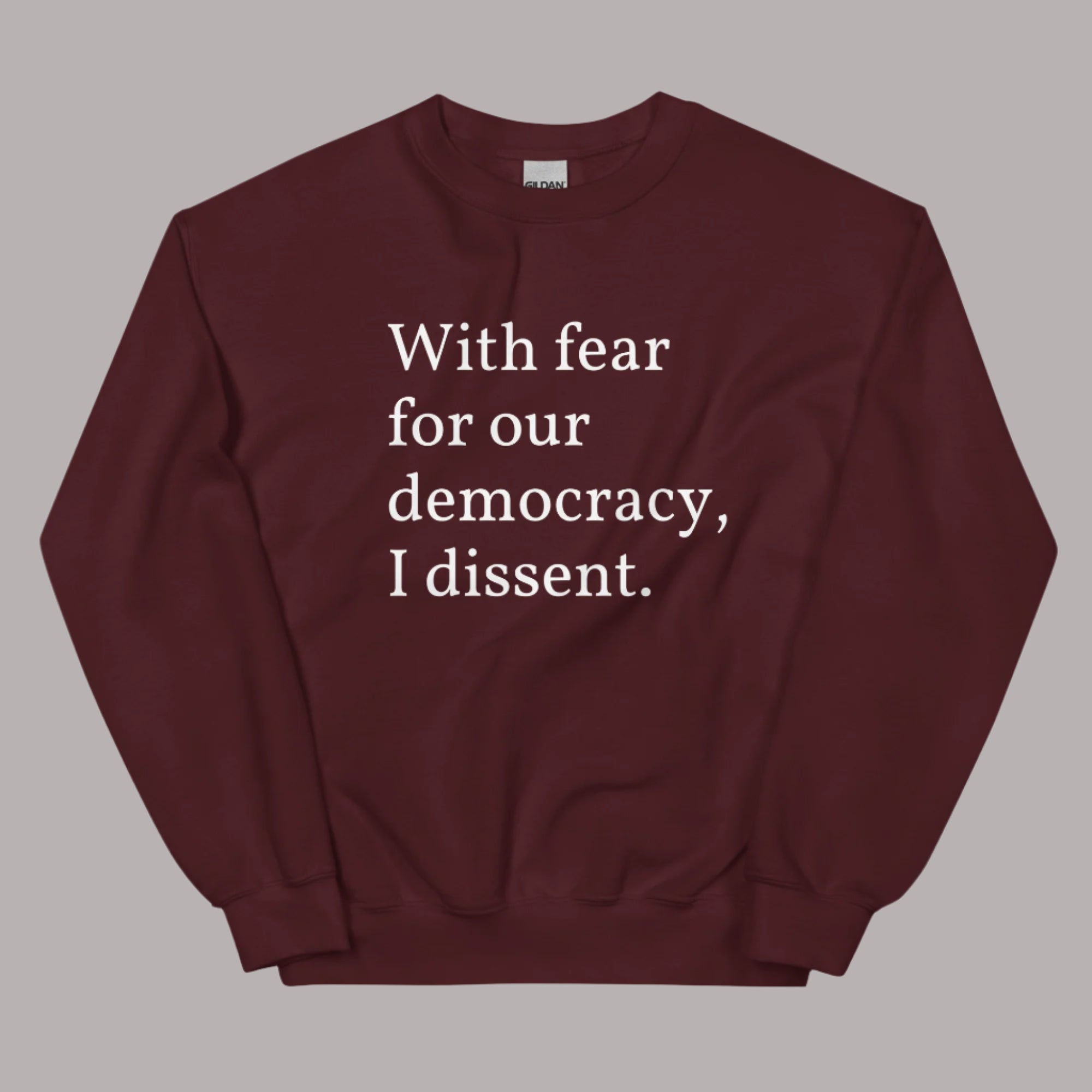 Flat lay of a maroon sweatshirt on a neutral background. The sweatshirt features white serif text across the chest that reads: “With fear for our democracy, I dissent.”
