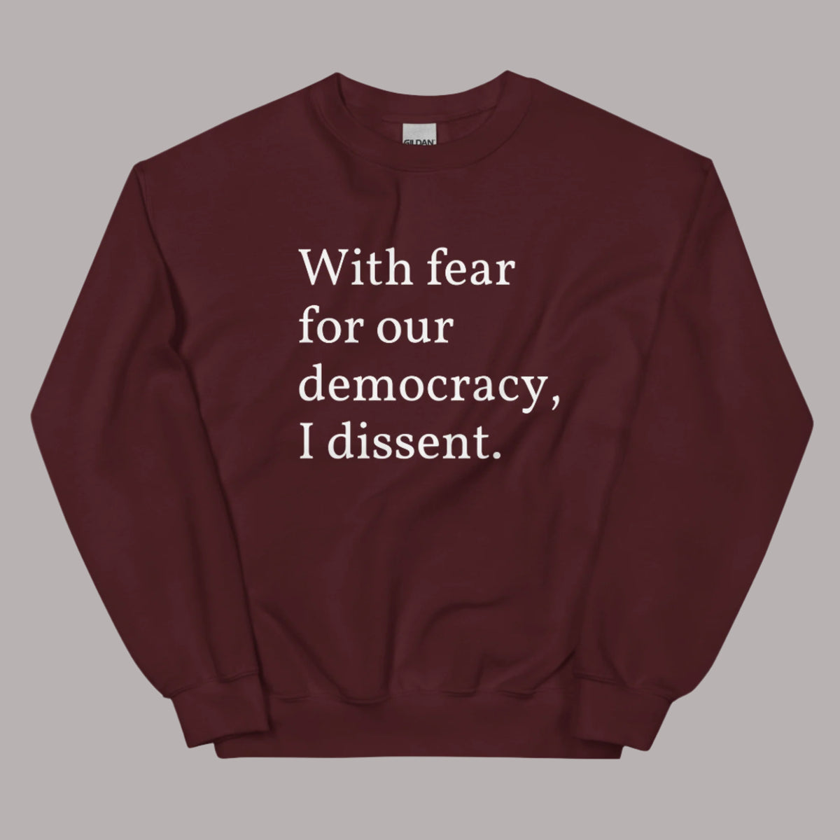 Flat lay of a maroon sweatshirt on a neutral background. The sweatshirt features white serif text across the chest that reads: “With fear for our democracy, I dissent.”