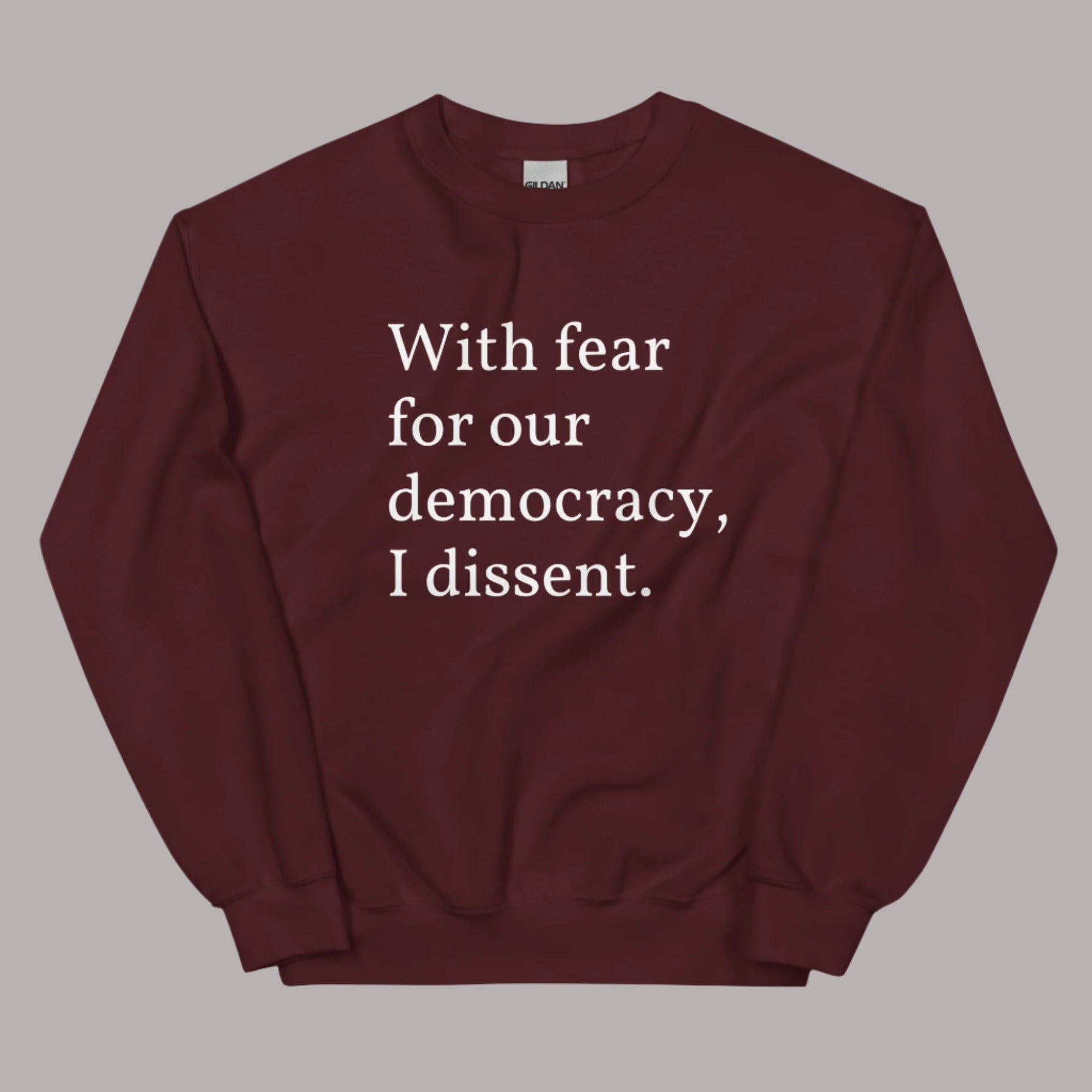 Flat lay of a maroon sweatshirt on a neutral background. The sweatshirt features white serif text across the chest that reads: “With fear for our democracy, I dissent.”