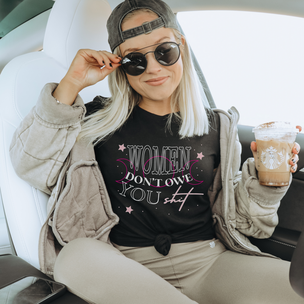 Woman holds a coffee wearing sunglasses, a ball cap, and a shirt that reads "women don't owe you shit."