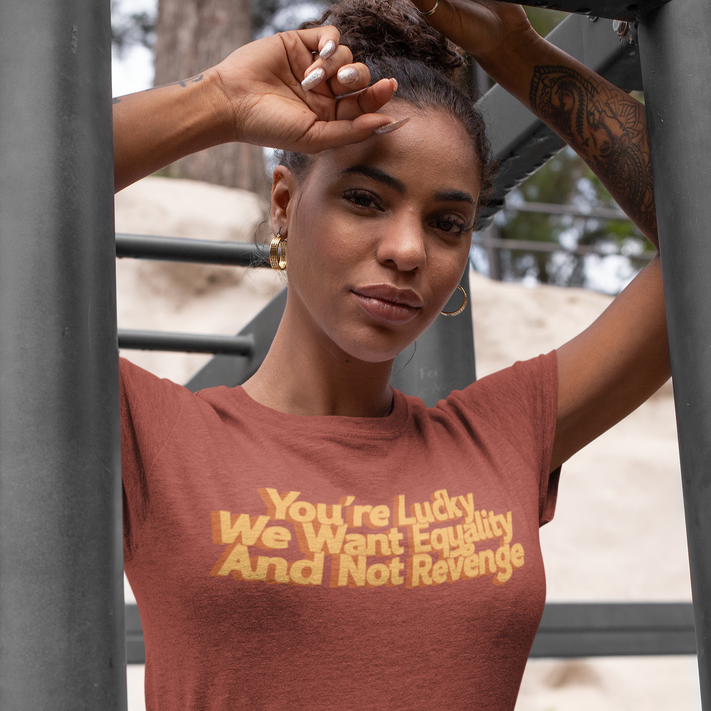 Close-up of a woman standing outdoors wearing a triblend unisex clay-colored T-shirt. The shirt features vintage gold text that reads, “you’re lucky we want equality and not revenge.” The soft fabric and subtle earth tone contrast with the striking message, making it a powerful and stylish statement piece.