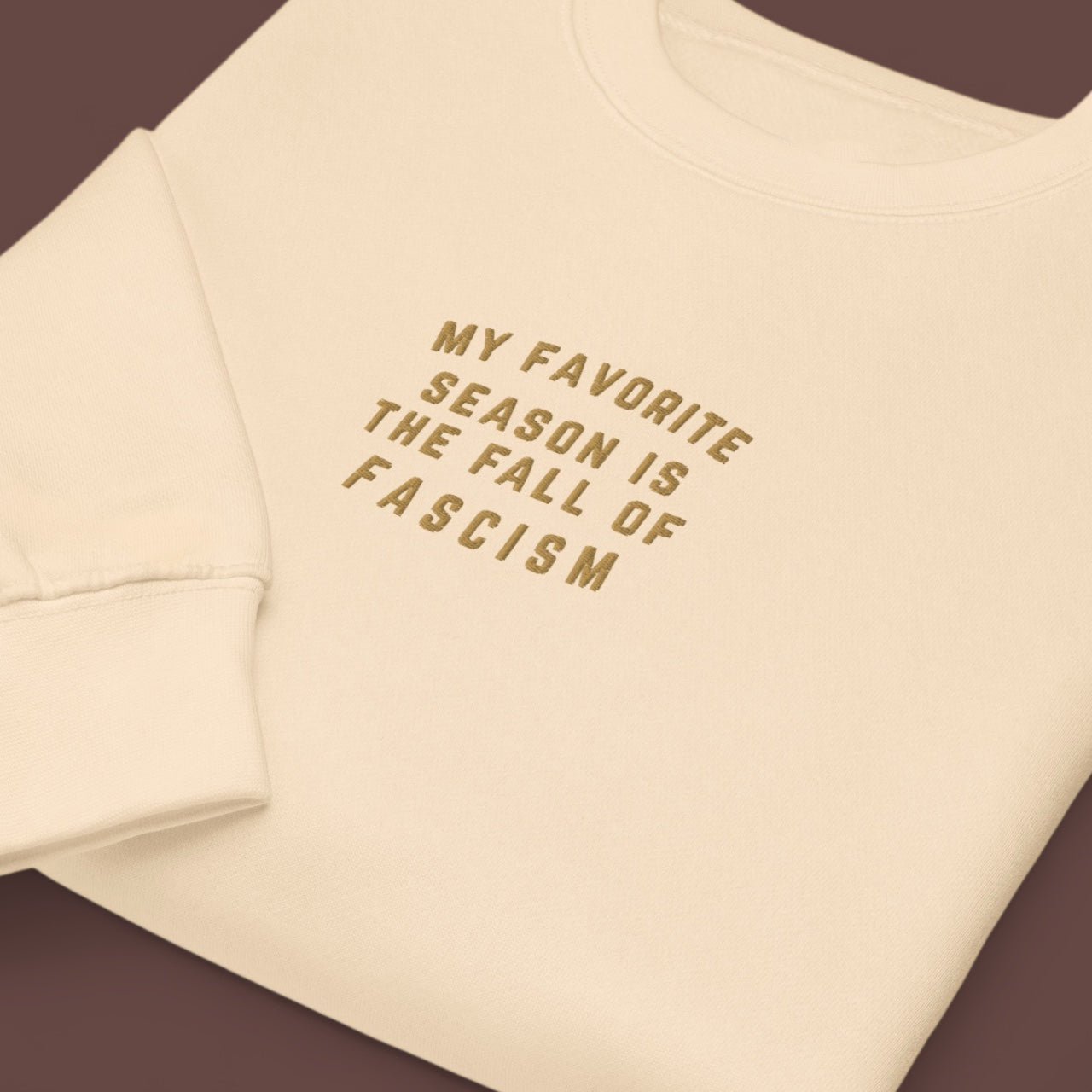 Beige sweatshirt with embroidered text 'My favorite season is the fall of fascism' on a brown background