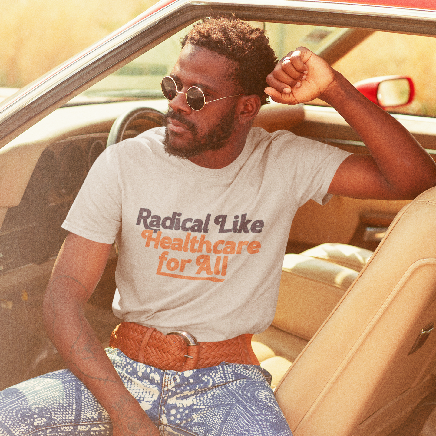 A man sits in a retro car wearing a heather dust-colored t-shirt that reads “Radical Like Healthcare for All” in a vintage-inspired font. The shirt has a soft, worn-in look that complements the classic vibe of the scene.