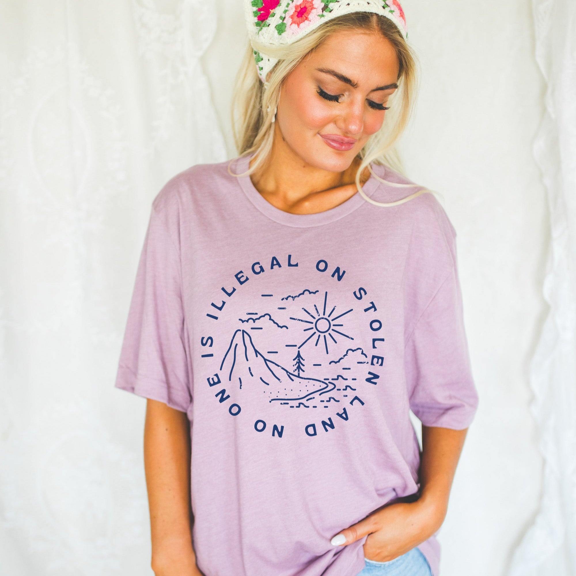 Woman wearing a purple t-shirt with a graphic design and text that reads "no one is illegal on stolen land" standing against a plain background.