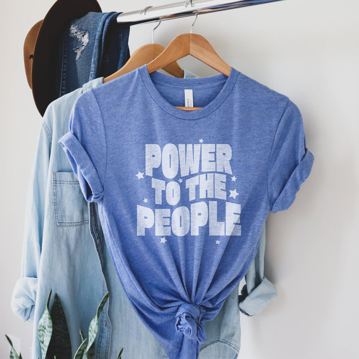 A light blue triblend t shirt hanging on a rack styled with a jean shirt and hat. The white font is faded and reads "power to the people" with white stars.