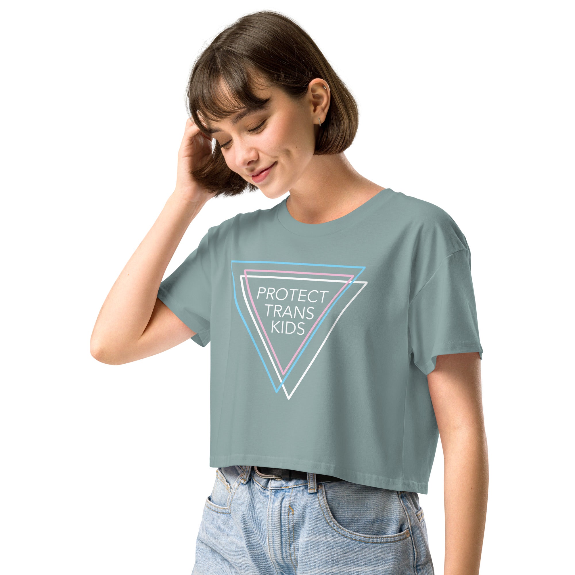 A woman standing confidently wearing a blue crop top that reads "Protect Trans Kids" in bold white letters, accented by three triangles in blue, pink, and white—the colors of the transgender flag. The crop top has a relaxed fit with dropped shoulders and a cropped length, styled with high-waisted jeans.
