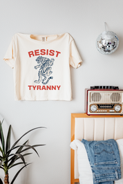 Beige t-shirt with 'Resist Tyranny' design on a wall above a bed with denim jacket.