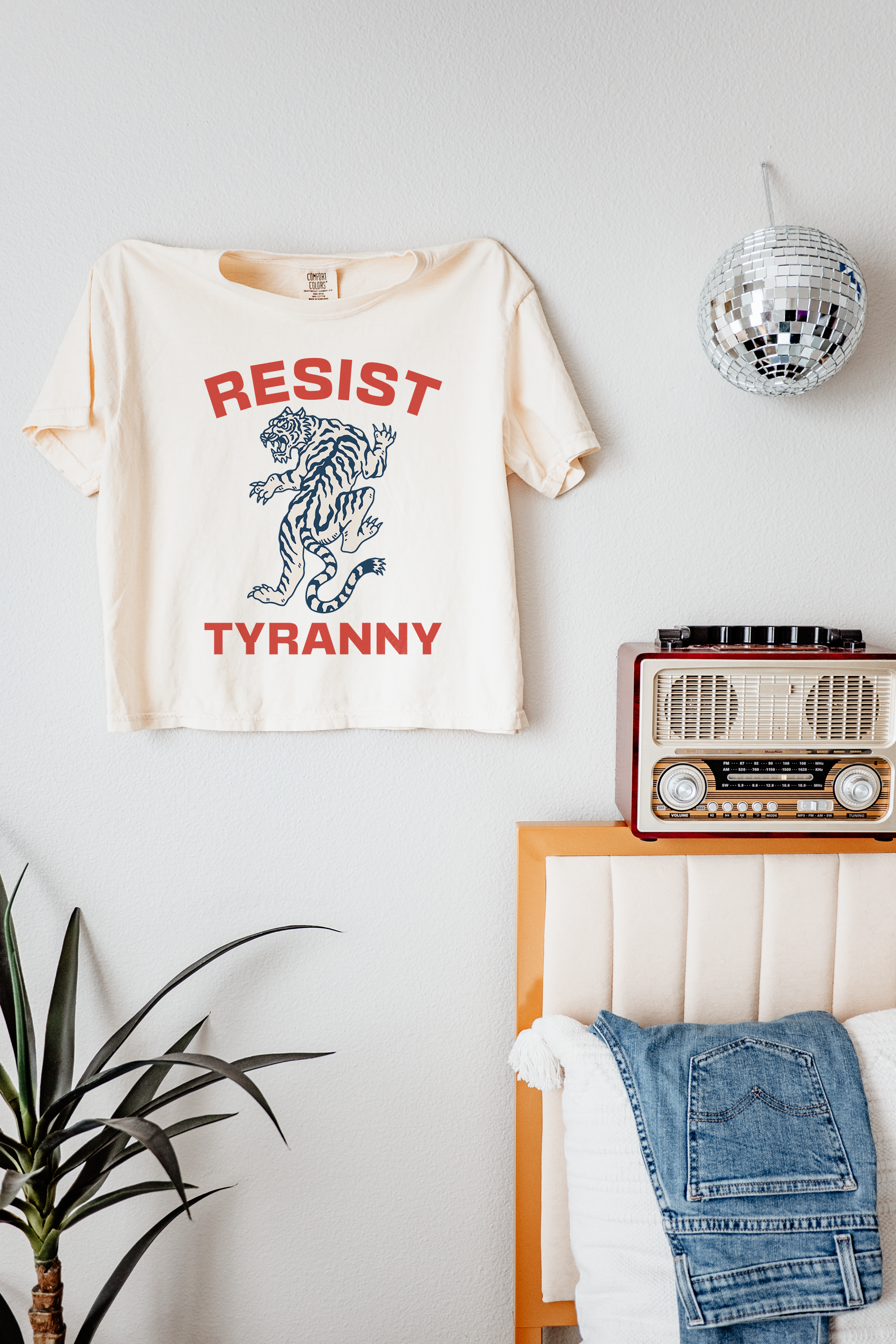 Beige t-shirt with 'Resist Tyranny' design on a wall above a bed with denim jacket.